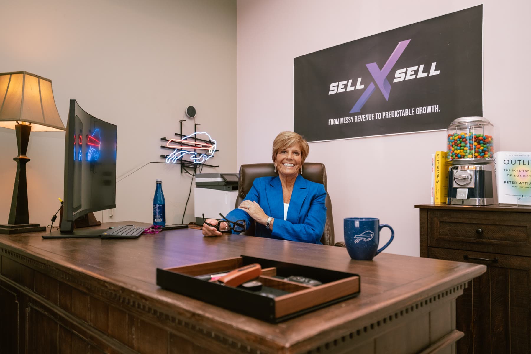 Shelley Dunagan seated at her desk in a blue blazer with the SellXSell sign behind her.